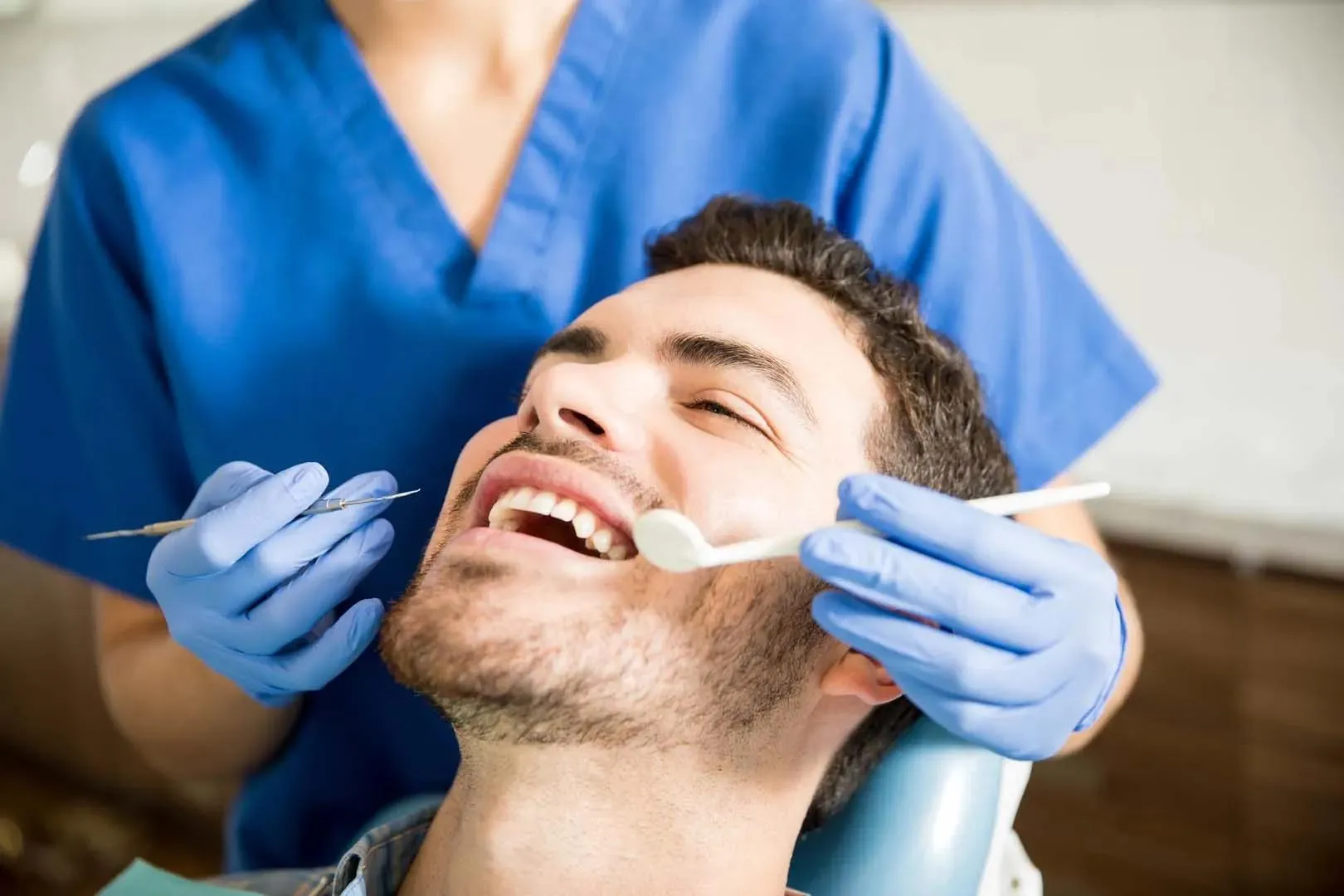 Dentist wearing blue gloves examining a smiling man's teeth with dental tools.