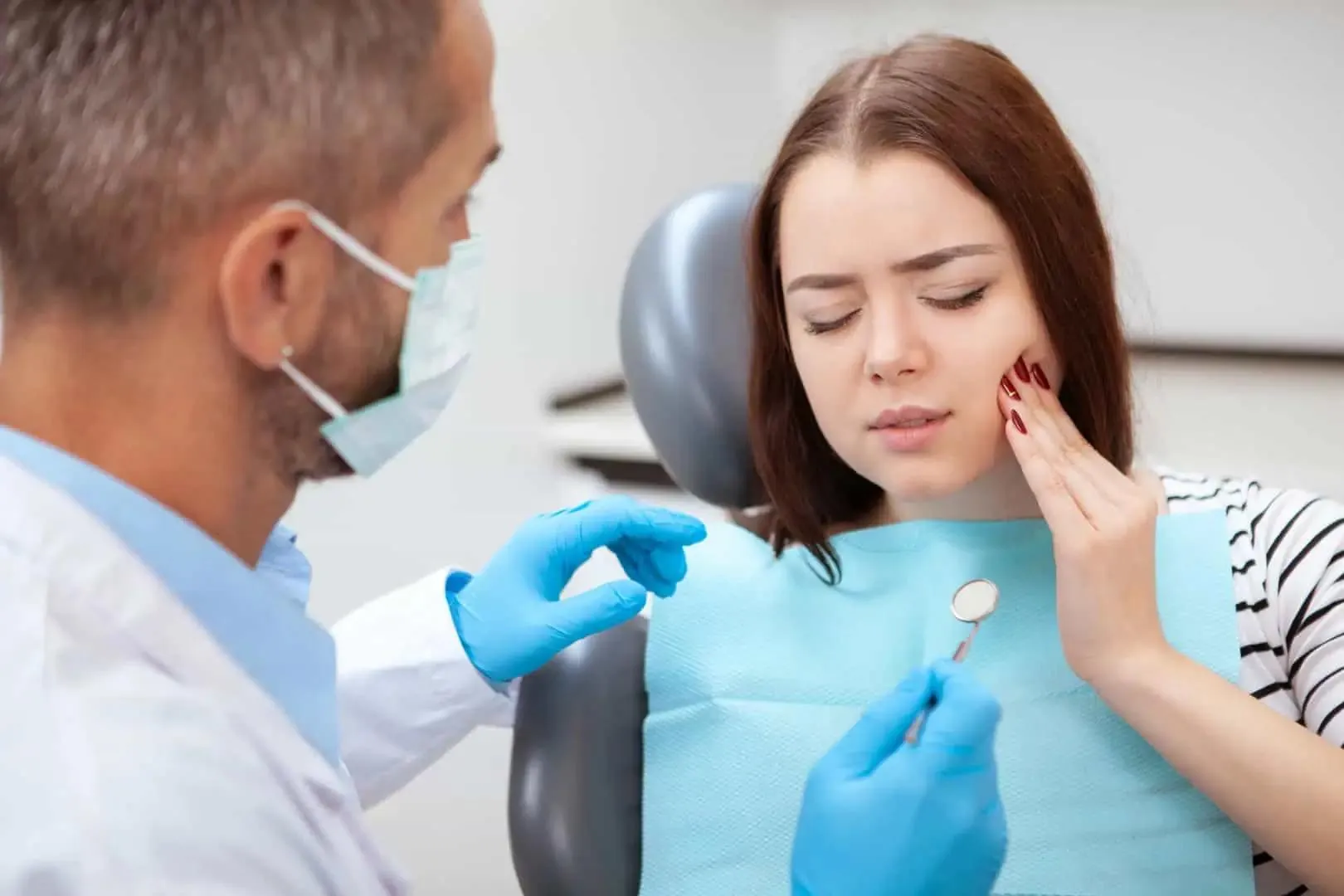 Young woman wincing in dental chair touches her jaw as dentist with gloves and mask holds dental mirror.