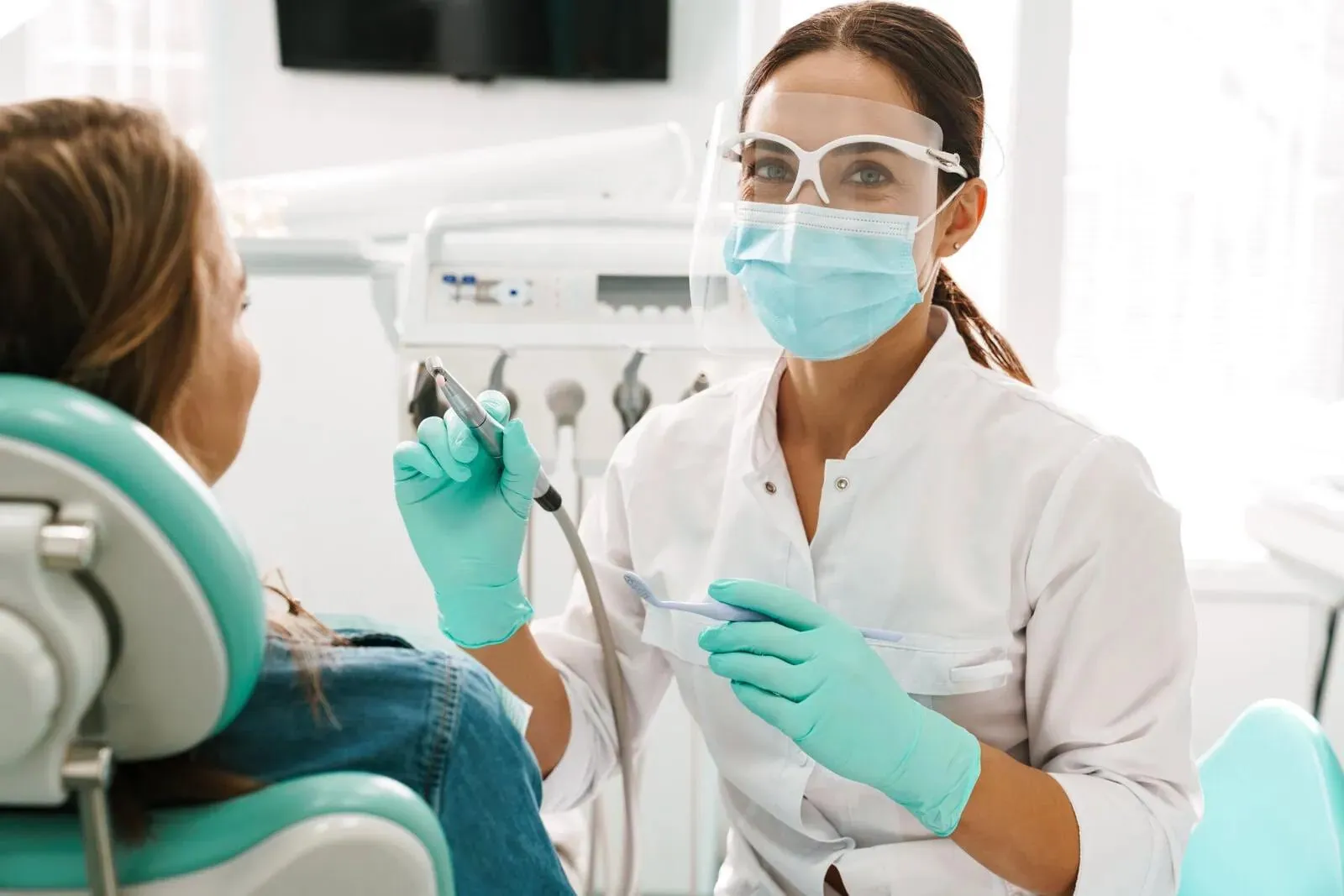 Dentist wearing mask, gloves, and face shield holding dental tools with patient seated in chair.