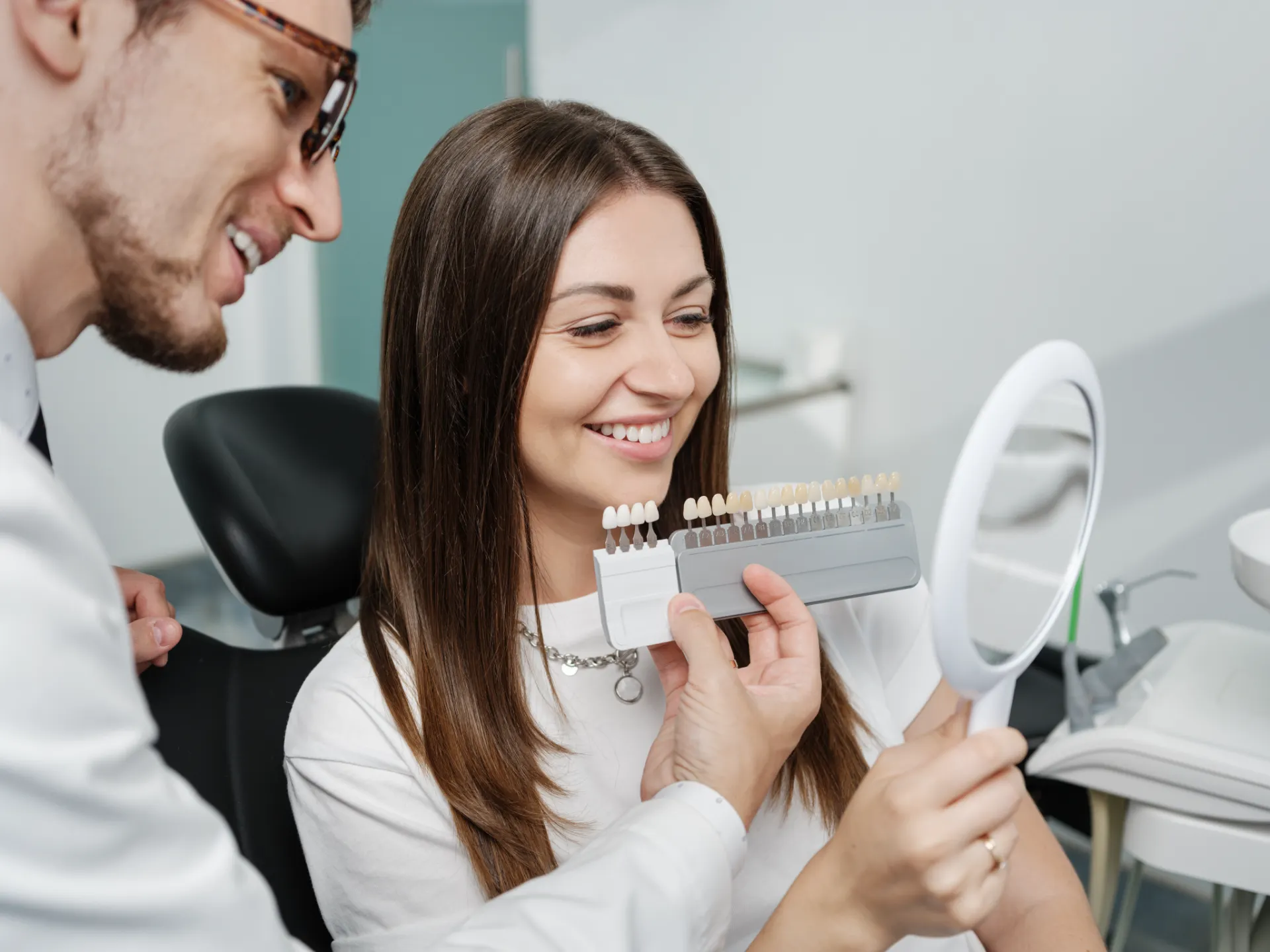 Dentist showing a female patient different tooth shades as she smiles holding a mirror.