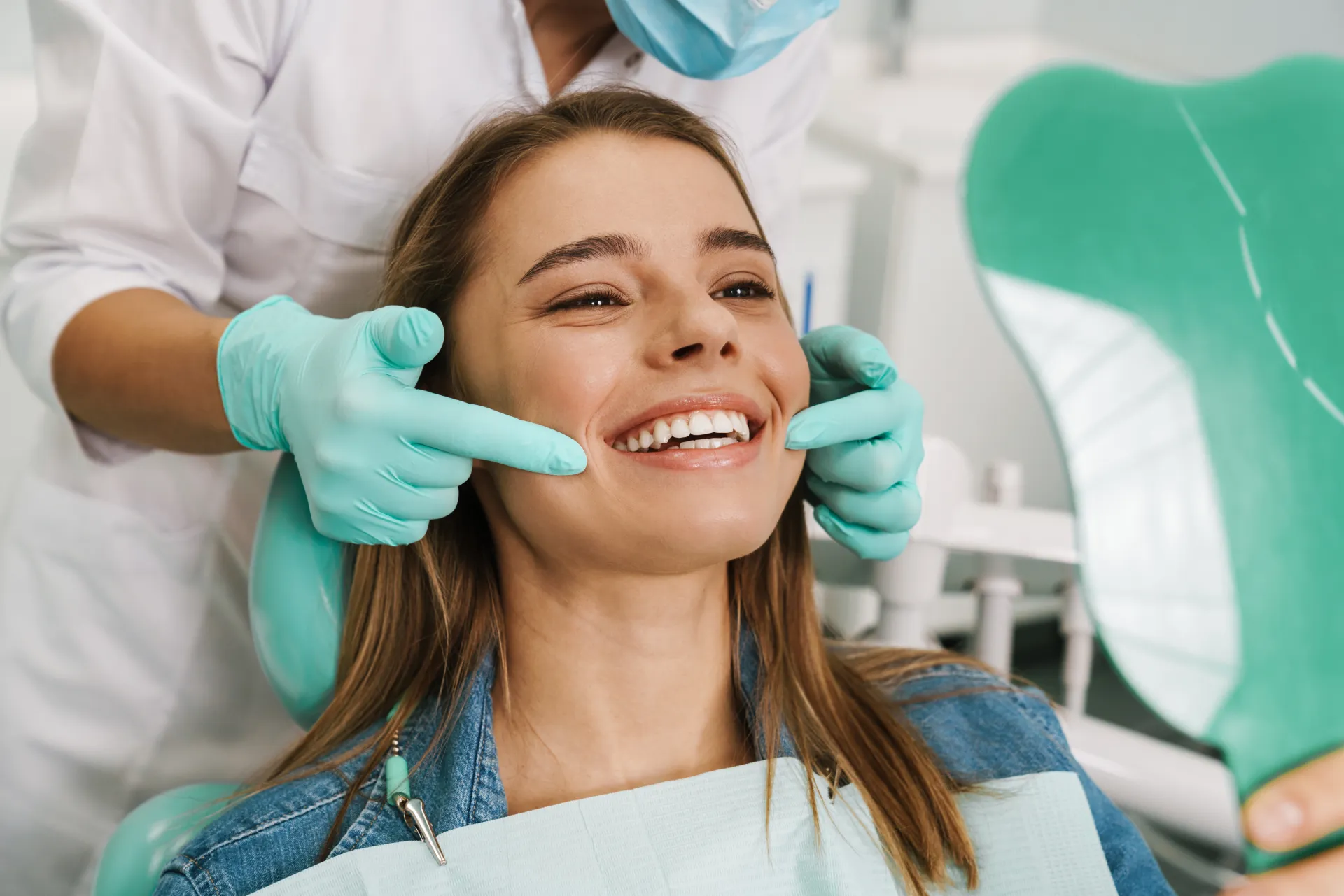 Dentist wearing gloves examines smiling woman's teeth as she looks at a dental mirror in clinic.