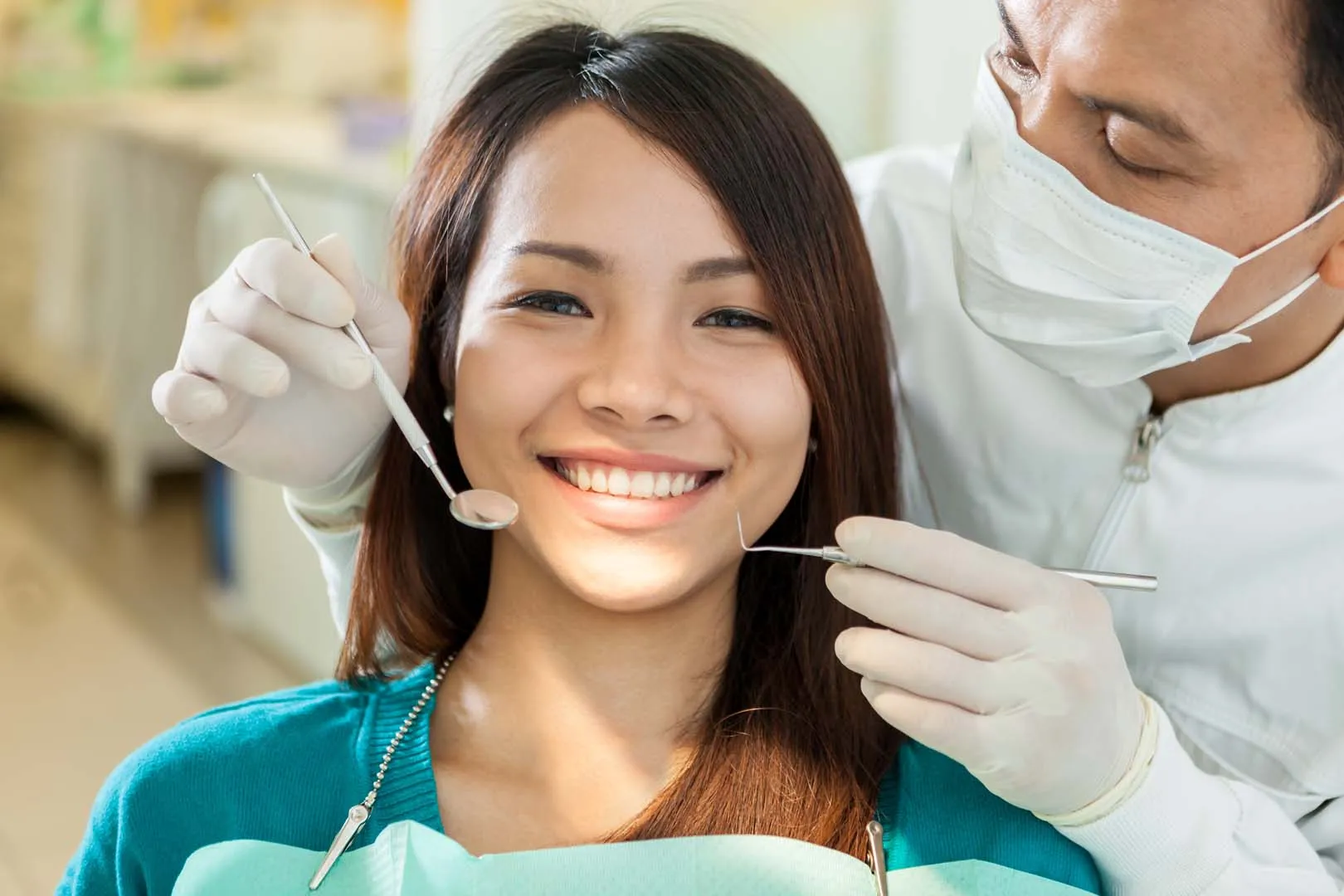 Dentist in mask examining smiling woman's teeth using dental mirror and probe.